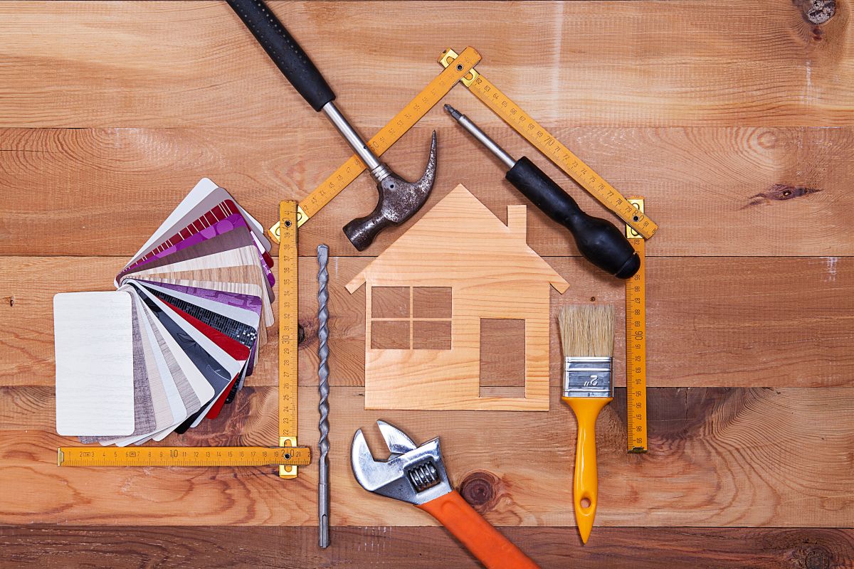 Overhead arrangement of hand tools, rulers, paint swatches, and a wooden house-shaped piece on a wooden work surface.