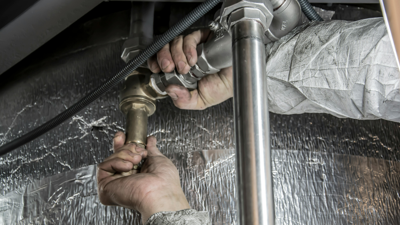 Close-up of gloved hands connecting metal plumbing pipes beneath a sink or fixture, lit with bright indoor lighting.