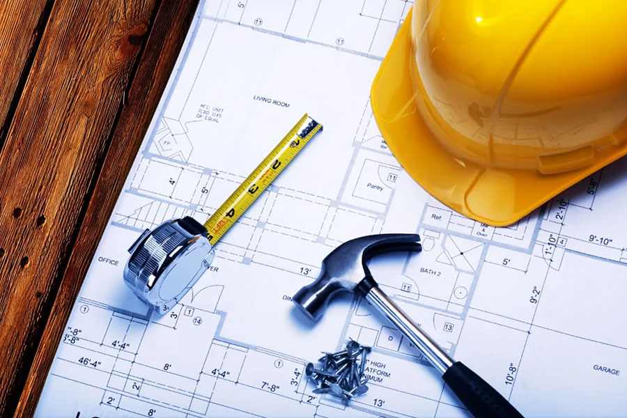 Overhead view of a yellow hard hat, tape measure, hammer, and building plans on a wooden surface, brightly lit.